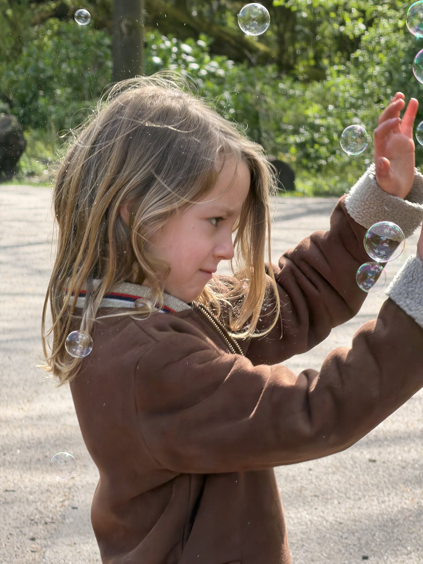 A young child with blonde hair, wearing a brown jacket, is captured mid-action, reaching out to interact with numerous floating bubbles